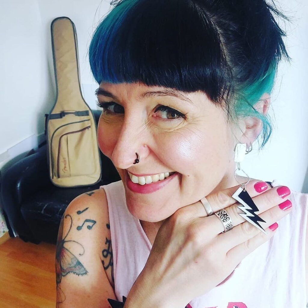 Eine Frau präsentiert die Ohrhänger mit einem Blitz auf iherre Hand. Die Nägel sind pink lackiert, sie trägt Tattoos, im Hintergrund steht eine Gitarrentasche auf einem Sofa. Die Szene ist privat.