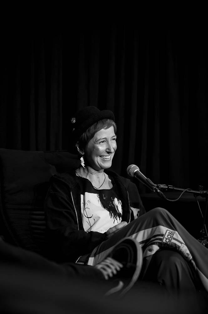 Eine Frau mit Kurzhaarschnitt und Hut sitzt in ihrer Podcastveranstaltung bei einer LiveAufnahme. An ihrem Ohr ist der Blitzohrhänger zu sehen, sie trägt die matt-silberne Seite nach außen, die eloxierte schwarze Seite innen ist nicht sichtbar.