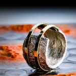 Das Bild zeigt einen Silber-Ring, dessen Ringkopf offen ist und der eine gebrannte Oberfläche hat. Er ird von zwei gehämmerten Goldringen mit polierter Oberfläche zusammengehalten. Der Hintergrund zeigt eine unebene anthrazitfarbene Fläche mit leuchtenden Farbakzenten in Orange.