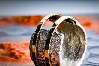 Das Bild zeigt einen Silber-Ring, dessen Ringkopf offen ist und der eine gebrannte Oberfläche hat. Er ird von zwei gehämmerten Goldringen mit polierter Oberfläche zusammengehalten. Der Hintergrund zeigt eine unebene anthrazitfarbene Fläche mit leuchtenden Farbakzenten in Orange.