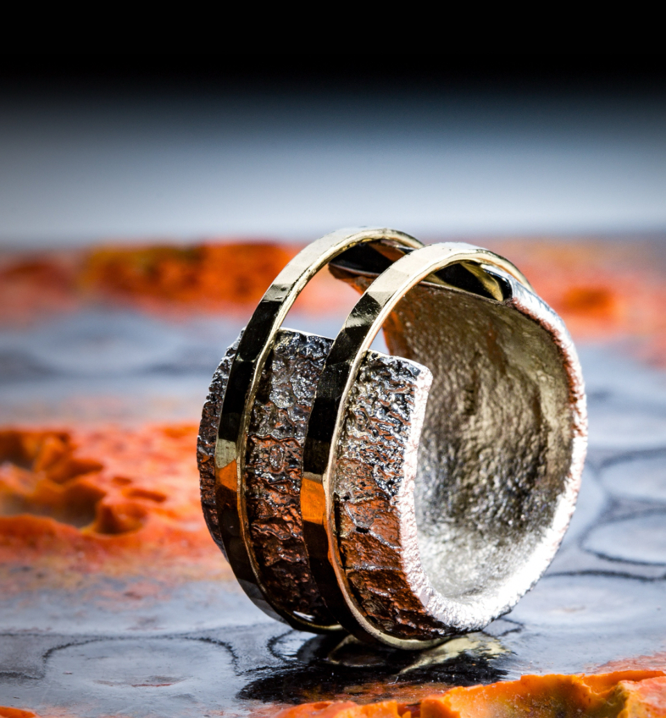 Das Bild zeigt einen Silber-Ring, dessen Ringkopf offen ist und der eine gebrannte Oberfläche hat. Er ird von zwei gehämmerten Goldringen mit polierter Oberfläche zusammengehalten. Der Hintergrund zeigt eine unebene anthrazitfarbene Fläche mit leuchtenden Farbakzenten in Orange.