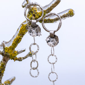 Auf dem Bild siehst du lange Ohrhänger mit einem unregelmäßig geformten Ring, einem gewölbten Silberteil mit einem Stern, vintage geschwärtzt, mit gehämmerter grober Kette. Der Ohrringständer ist ein St in der Natur. Er ist von einer Eisschicht überzogen.