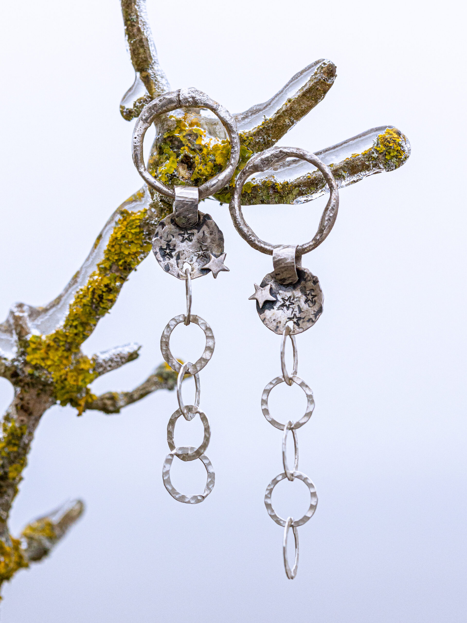 Auf dem Bild siehst du lange Ohrhänger mit einem unregelmäßig geformten Ring, einem gewölbten Silberteil mit einem Stern, vintage geschwärtzt, mit gehämmerter grober Kette. Der Ohrringständer ist ein St in der Natur. Er ist von einer Eisschicht überzogen.