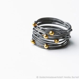 Auf dem Bild ist ein geschwärzter Wickelring aus geschlungenem Silberdraht zu sehen, auf den sechs Goldkugeln ungleichmäßig im Bereich des Ringkopfs verteilt sind.