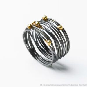 Auf dem Bild ist ein geschwärzter Wickelring aus geschlungenem Silberdraht zu sehen, auf den sechs Goldkugeln ungleichmäßig im Bereich des Ringkopfs verteilt sind.