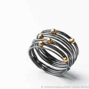 Auf dem Bild ist ein geschwärzter Wickelring aus geschlungenem Silberdraht zu sehen, auf den sechs Goldkugeln ungleichmäßig im Bereich des Ringkopfs verteilt sind.