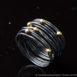 Auf dem Bild ist ein geschwärzter Wickelring aus geschlungenem Silberdraht zu sehen, auf den sechs Goldkugeln ungleichmäßig im Bereich des Ringkopfs verteilt sind.