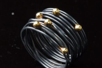 Auf dem Bild ist ein geschwärzter Wickelring aus geschlungenem Silberdraht zu sehen, auf den sechs Goldkugeln ungleichmäßig im Bereich des Ringkopfs verteilt sind.