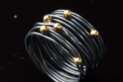 Auf dem Bild ist ein geschwärzter Wickelring aus geschlungenem Silberdraht zu sehen, auf den sechs Goldkugeln ungleichmäßig im Bereich des Ringkopfs verteilt sind.