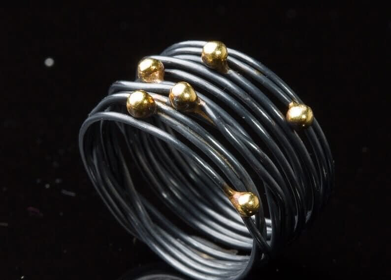 Auf dem Bild ist ein geschwärzter Wickelring aus geschlungenem Silberdraht zu sehen, auf den sechs Goldkugeln ungleichmäßig im Bereich des Ringkopfs verteilt sind.