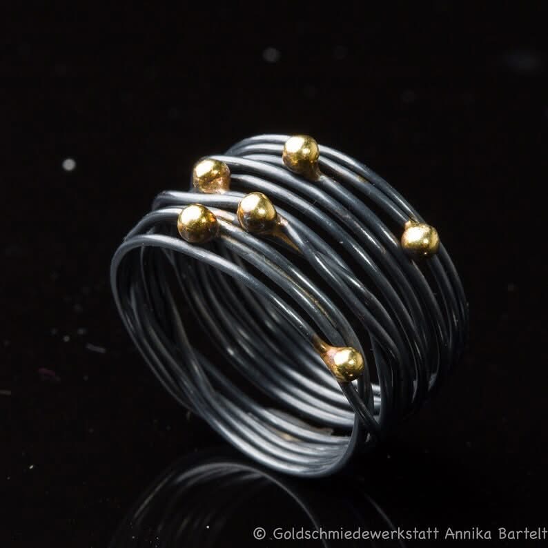 Auf dem Bild ist ein geschwärzter Wickelring aus geschlungenem Silberdraht zu sehen, auf den sechs Goldkugeln ungleichmäßig im Bereich des Ringkopfs verteilt sind.