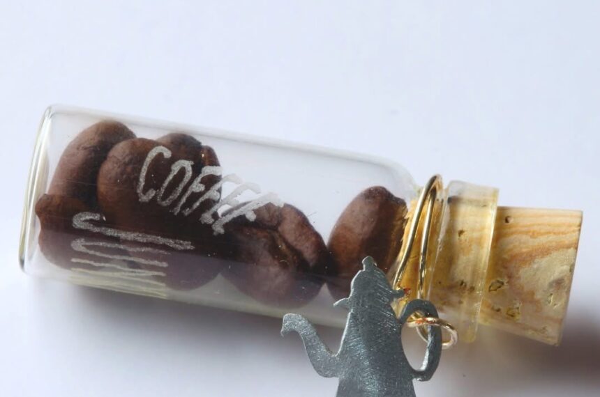Eine Phiole mit Kaffeebohnen, die eine Gravur hat. Text: Coffee Junky. Um den Korken ist eine Drahtspirale gebunden, an der eine kleine Kaffekanne als Anhänger aus Metall angebracht ist.