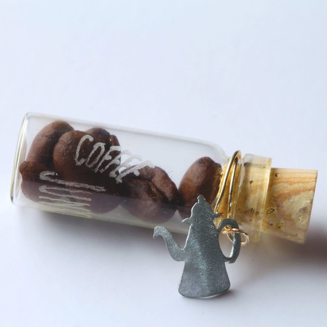 Eine Phiole mit Kaffeebohnen, die eine Gravur hat. Text: Coffee Junky. Um den Korken ist eine Drahtspirale gebunden, an der eine kleine Kaffekanne als Anhänger aus Metall angebracht ist.