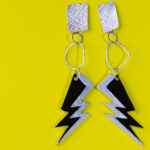 Ohrhänger mit Strukturierten Elementen in unregelmäßiger Form vor einem gelben Hintergrund. Den Abschluss bilden zwei Blitze aus schwarz eloxiertem Aluminium.