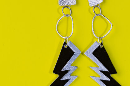 Ohrhänger mit Strukturierten Elementen in unregelmäßiger Form vor einem gelben Hintergrund. Den Abschluss bilden zwei Blitze aus schwarz eloxiertem Aluminium.