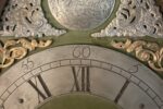 Das Bild zeigt den Ausschnitt eines gravierten Zifferblattes einer antiken Standuhr mit Stunden und Minuten. Darüber goldenen und silberne Ornamente und eine Gravurplatte