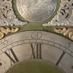 Das Bild zeigt den Ausschnitt eines gravierten Zifferblattes einer antiken Standuhr mit Stunden und Minuten. Darüber goldenen und silberne Ornamente und eine Gravurplatte