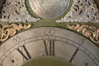 Das Bild zeigt den Ausschnitt eines gravierten Zifferblattes einer antiken Standuhr mit Stunden und Minuten. Darüber goldenen und silberne Ornamente und eine Gravurplatte