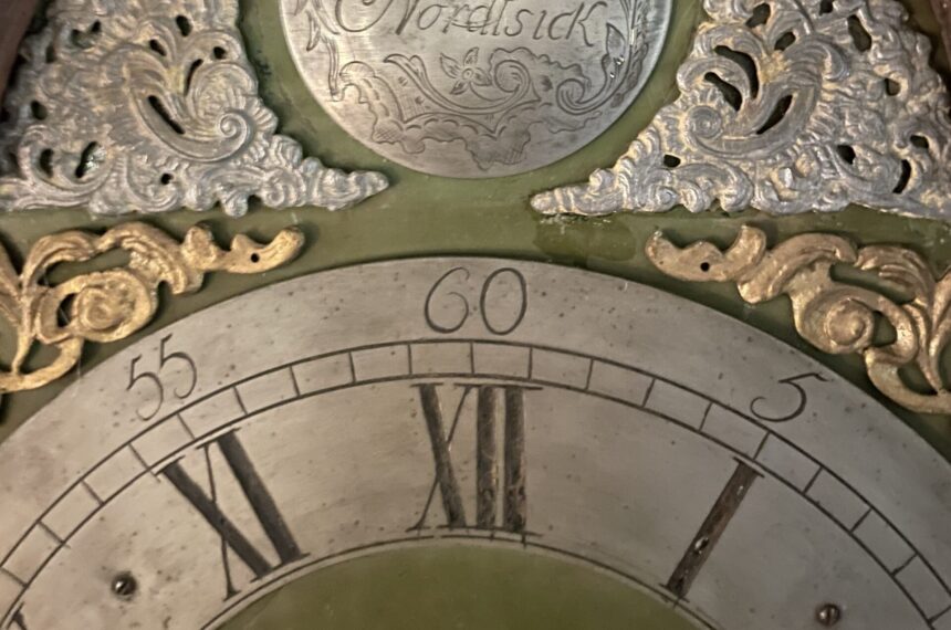 Das Bild zeigt den Ausschnitt eines gravierten Zifferblattes einer antiken Standuhr mit Stunden und Minuten. Darüber goldenen und silberne Ornamente und eine Gravurplatte