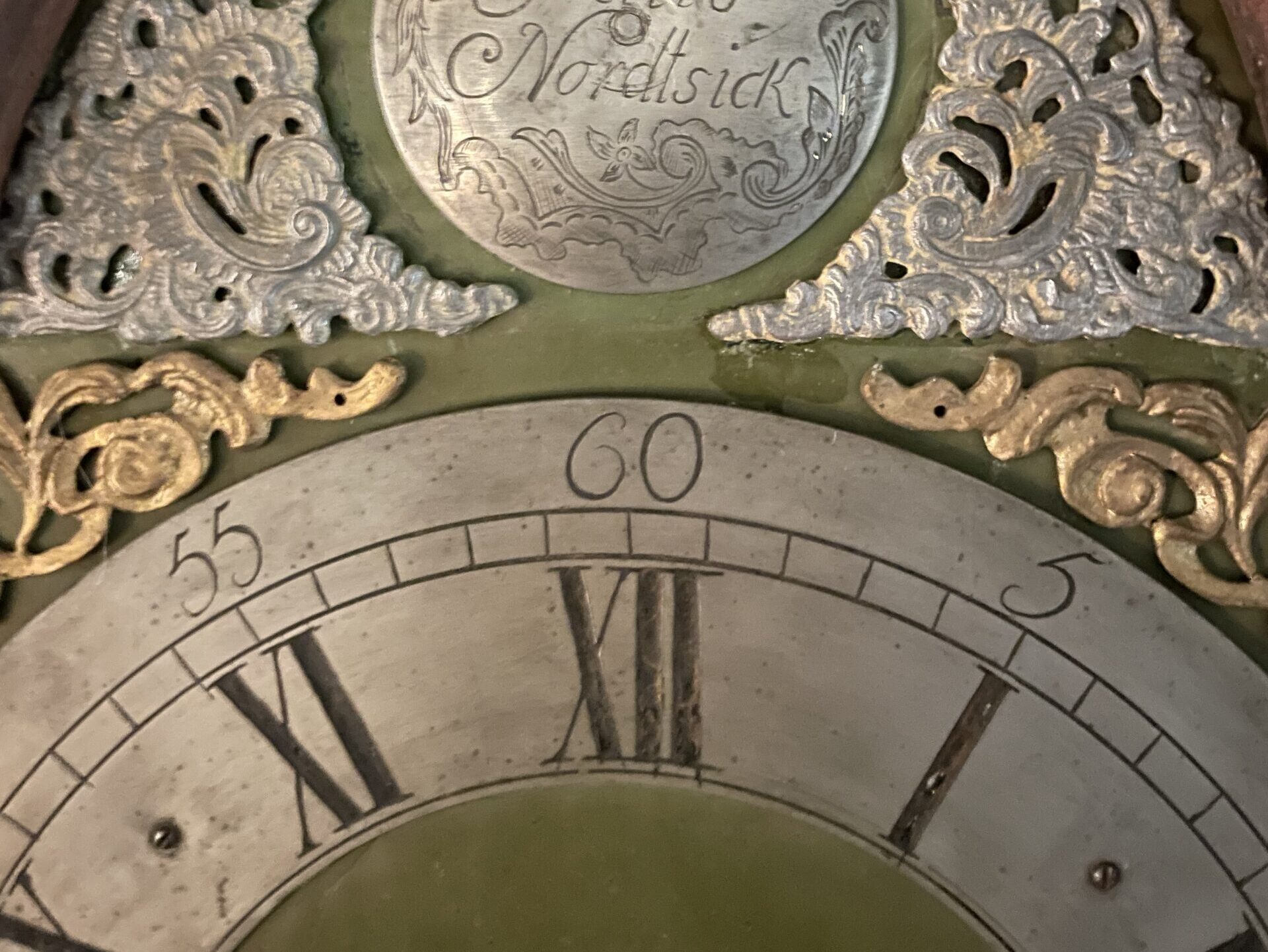 Das Bild zeigt den Ausschnitt eines gravierten Zifferblattes einer antiken Standuhr mit Stunden und Minuten. Darüber goldenen und silberne Ornamente und eine Gravurplatte